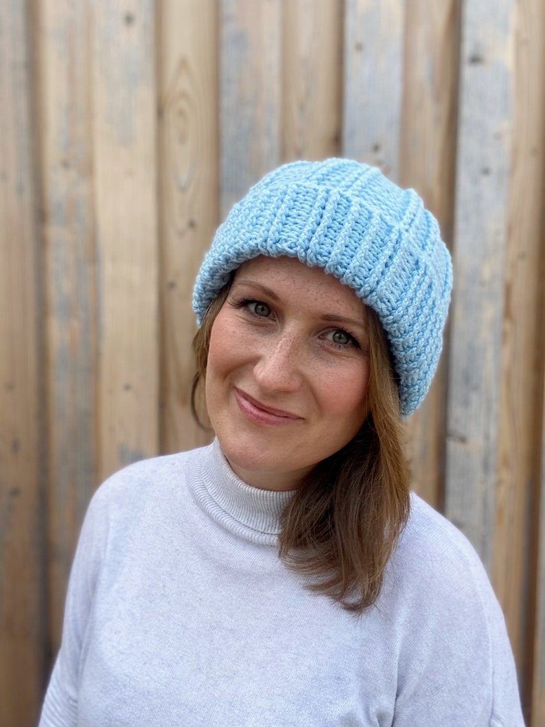 May include: A light blue knitted beanie hat with a ribbed texture. The hat is worn by a person with brown hair and is positioned on their head.