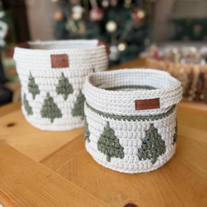 May include: Two cream-coloured crochet baskets featuring green Christmas tree patterns. Each basket has a brown leather tag. They are displayed on a wooden table. The larger basket is taller than the smaller one. The background is blurred.
