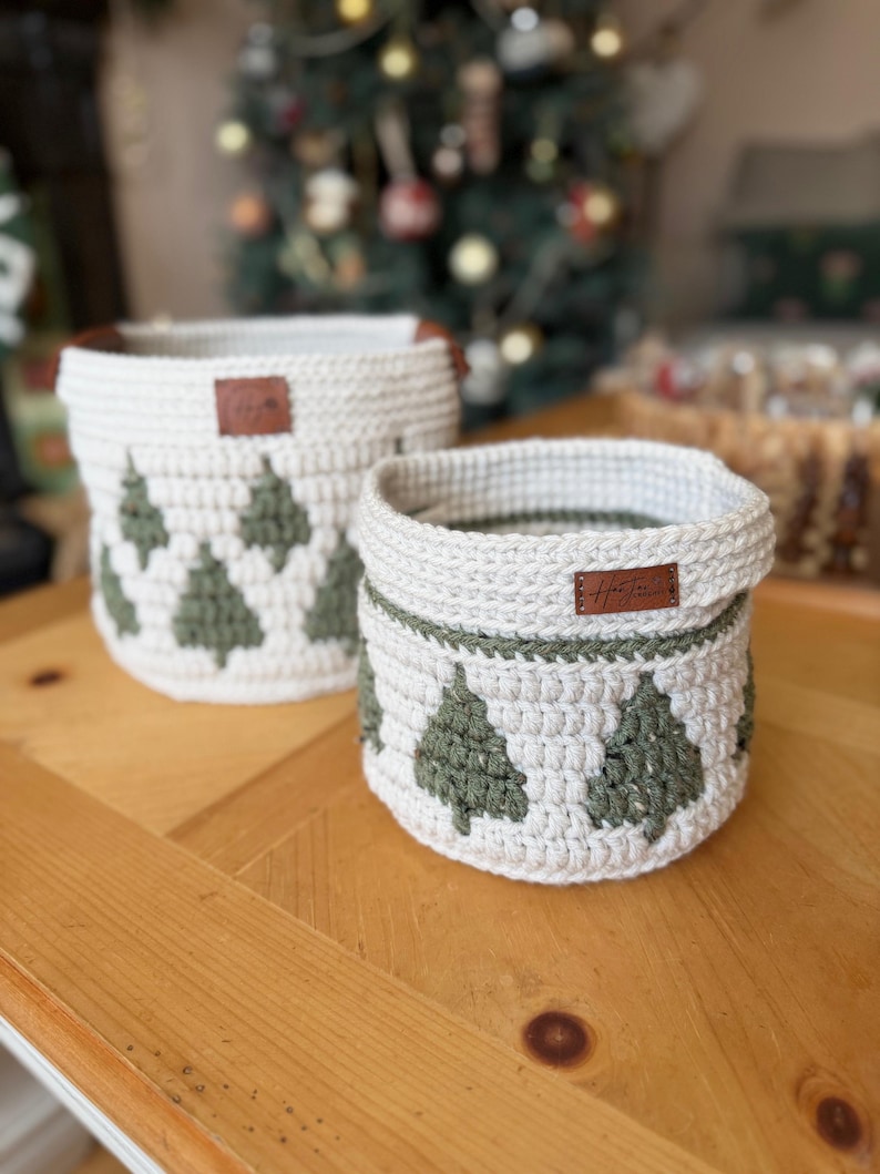 May include: Two cream-coloured crochet baskets featuring green Christmas tree patterns. Each basket has a brown leather tag. They are displayed on a wooden table. The larger basket is taller than the smaller one. The background is blurred.