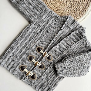 Pu&ograve; includere: Un cardigan di maglia grigia con bottoni marroni. Il cardigan ha una trama a coste ed &egrave; realizzato con un filato maculato.