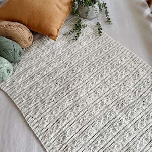 Peut inclure: Une couverture tricot&eacute;e gris clair avec un motif en point de c&acirc;ble. La couverture est pos&eacute;e sur une surface blanche, avec un coussin beige et une petite plante en pot en arri&egrave;re-plan. Plusieurs pelotes de laine vertes, beiges et blanches sont visibles.