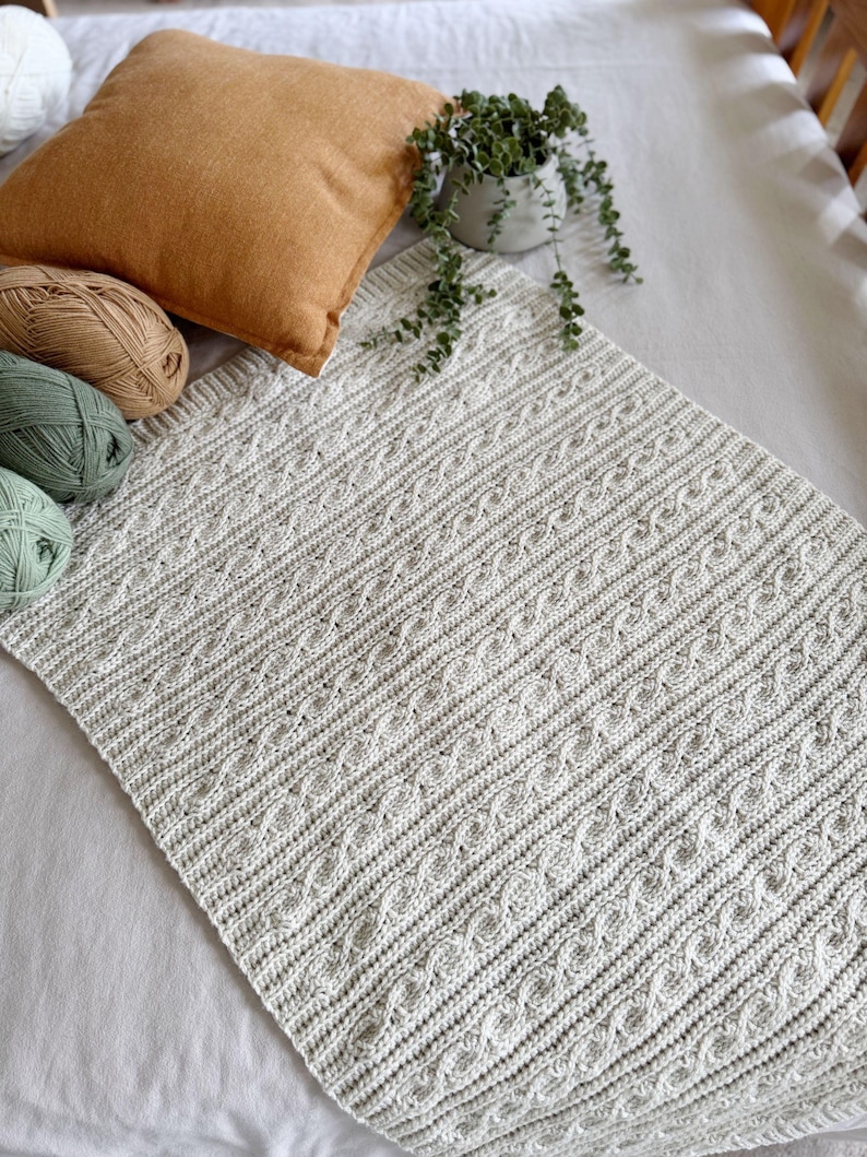 Peut inclure: Une couverture tricot&eacute;e gris clair avec un motif en point de c&acirc;ble. La couverture est pos&eacute;e sur une surface blanche, avec un coussin beige et une petite plante en pot en arri&egrave;re-plan. Plusieurs pelotes de laine vertes, beiges et blanches sont visibles.
