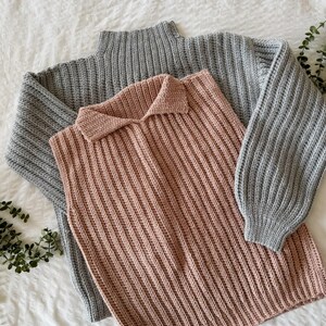 May include: Two knitted sweaters. One is a light grey, long-sleeved sweater with a ribbed texture. The other is a sleeveless, collared sweater in a dusty rose colour, also with a ribbed texture. Both are lying on a white surface.