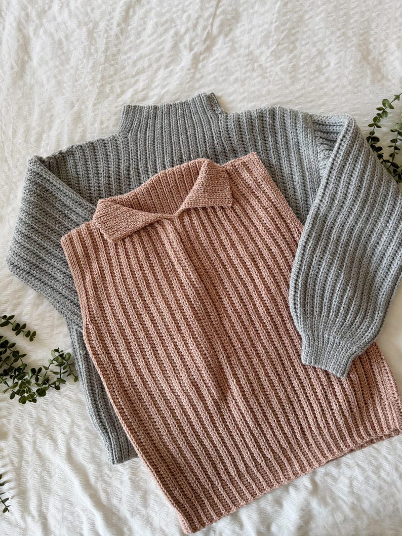 May include: Two knitted sweaters. One is a light grey, long-sleeved sweater with a ribbed texture. The other is a sleeveless, collared sweater in a dusty rose colour, also with a ribbed texture. Both are lying on a white surface.