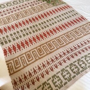 May include: A beige crocheted blanket with a geometric pattern in shades of brown, red, and green. The blanket features rows of diamonds, squares, and other shapes. The blanket is displayed on a white bed.