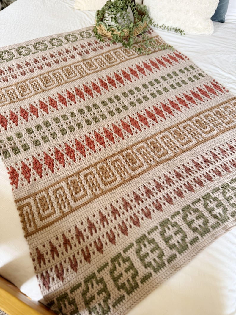 May include: A beige crocheted blanket with a geometric pattern in shades of brown, red, and green. The blanket features rows of diamonds, squares, and other shapes. The blanket is displayed on a white bed.