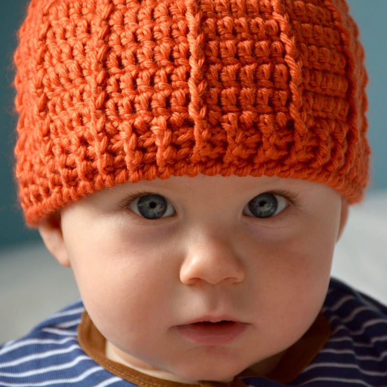 May include: A close-up of a baby wearing an orange crocheted hat. The baby has blue eyes and is looking directly at the camera.