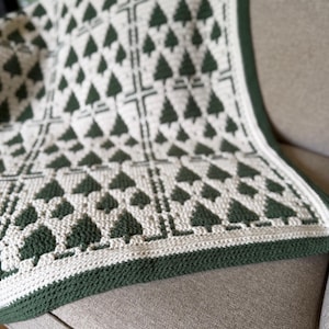 May include: A crochet blanket featuring a repeating pattern of dark green pine trees on an off-white background. The design is organized into rectangular sections separated by vertical and horizontal lines, finished with a solid dark green border.