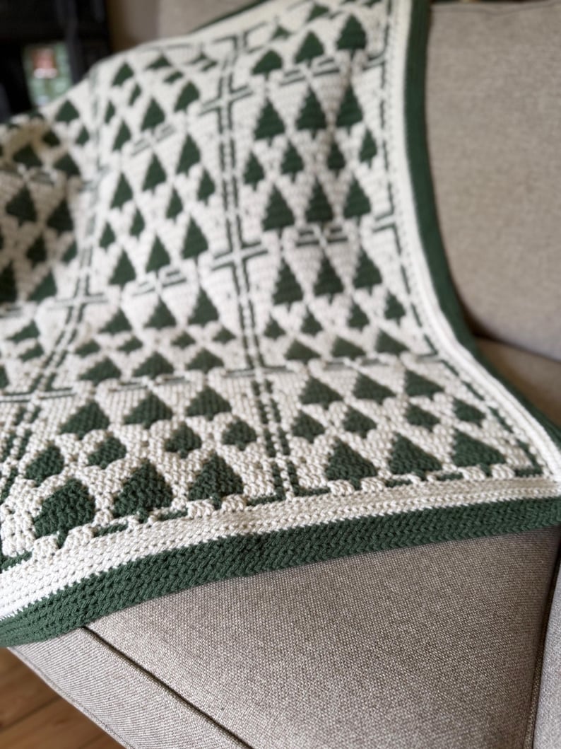 May include: A crochet blanket featuring a repeating pattern of dark green pine trees on an off-white background. The design is organized into rectangular sections separated by vertical and horizontal lines, finished with a solid dark green border.