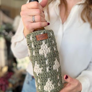 May include: A green and cream-colored crocheted wine bottle cozy. The cozy features a repeating tree pattern and a small brown leather tag with the word "Hunter" on it. The cozy is designed to fit around a standard wine bottle.