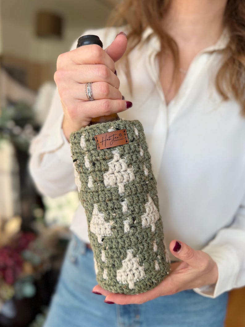 May include: A green and cream-colored crocheted wine bottle cozy. The cozy features a repeating tree pattern and a small brown leather tag with the word "Hunter" on it. The cozy is designed to fit around a standard wine bottle.