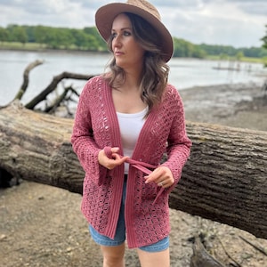 May include: A dusty rose crochet cardigan with long sleeves and a tie closure. The open-weave design is visible. The cardigan is worn over a white top and blue denim shorts. A straw hat with a wide brim is also worn.