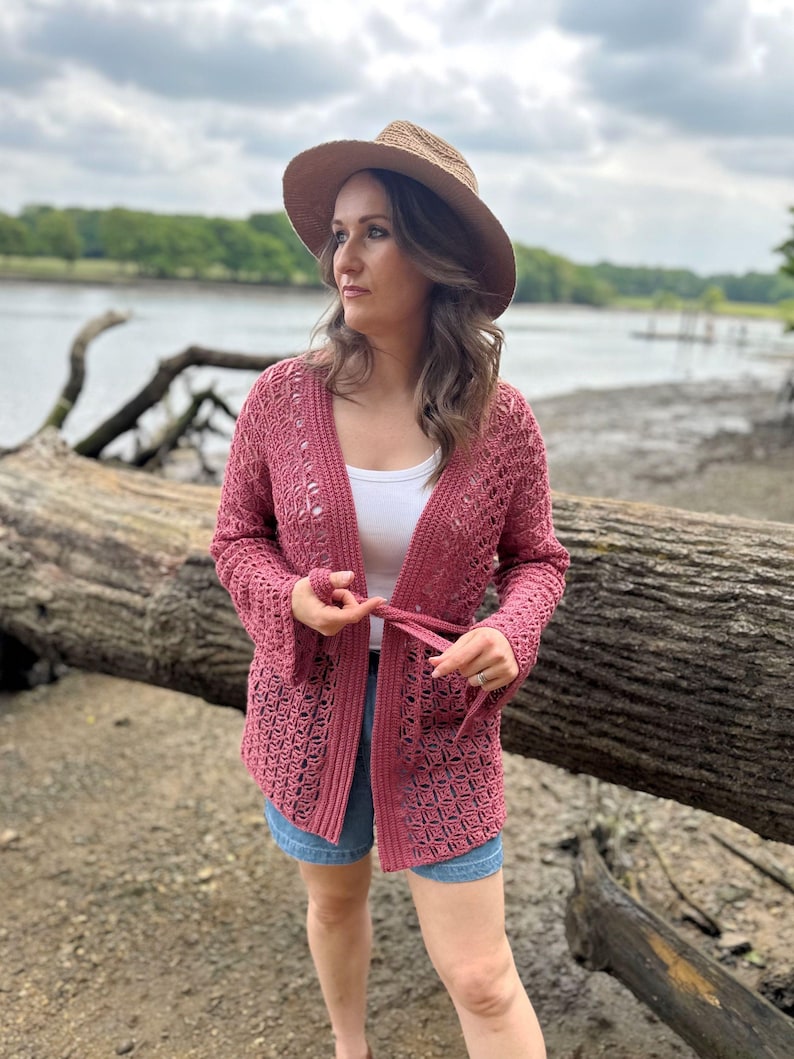 May include: A dusty rose crochet cardigan with long sleeves and a tie closure. The open-weave design is visible. The cardigan is worn over a white top and blue denim shorts. A straw hat with a wide brim is also worn.