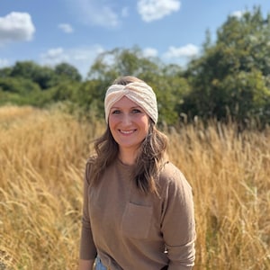 May include: A cream-colored, knitted headband with a twisted knot design. The headband is worn by a person in a field of tall grass. The person is wearing a brown long-sleeve shirt and blue jeans.