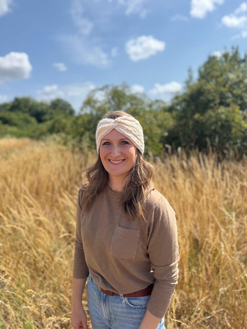 May include: A cream-colored, knitted headband with a twisted knot design. The headband is worn by a person in a field of tall grass. The person is wearing a brown long-sleeve shirt and blue jeans.