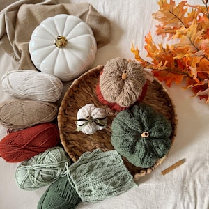 May include: An autumnal display featuring a white ceramic pumpkin, crocheted pumpkins in shades of brown and green, and skeins of yarn. A woven basket, autumn leaves, and a cinnamon stick add to the fall theme. The scene is set on a white surface.