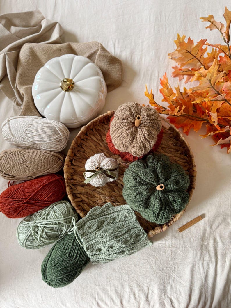 May include: An autumnal display featuring a white ceramic pumpkin, crocheted pumpkins in shades of brown and green, and skeins of yarn. A woven basket, autumn leaves, and a cinnamon stick add to the fall theme. The scene is set on a white surface.