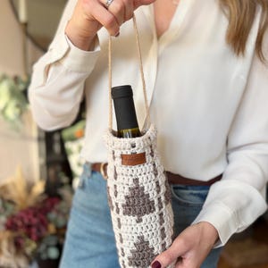 May include: A crocheted wine bottle holder in cream and brown, featuring a geometric pattern. The holder has a jute handle and a small leather tag. A wine bottle is inside the holder.