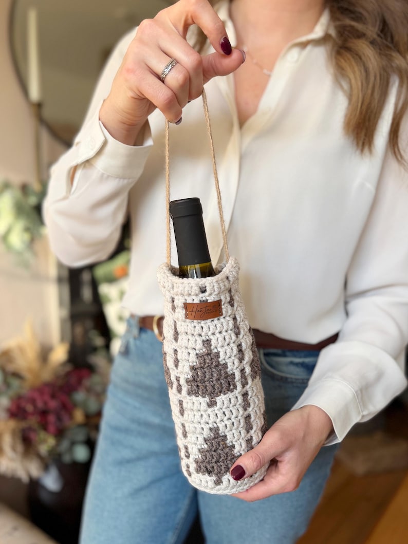 May include: A crocheted wine bottle holder in cream and brown, featuring a geometric pattern. The holder has a jute handle and a small leather tag. A wine bottle is inside the holder.