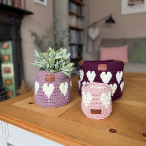 May include: Three crocheted heart-patterned baskets in varying sizes and shades of purple and pink. The baskets are arranged on a wooden table, with a plant in the largest basket. Each basket has a small leather tag.