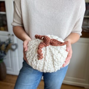 Peut inclure: Une citrouille blanche en crochet avec une tige en crochet marron. La citrouille est tenue par une personne portant un jean bleu.