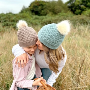 May include: Two knitted beanies with faux fur pom-poms. One is light brown, the other is light blue. A person is wearing the light blue beanie. The other person is wearing the light brown beanie. Both are in a field of tall grass.