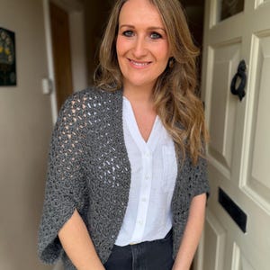 May include: A gray crochet shrug with openwork design. The shrug is worn over a white button-down shirt and black jeans. The outfit is casual and comfortable, suitable for layering.