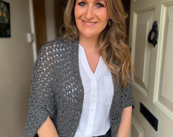 May include: A grey crochet shrug with an openwork pattern. The shrug is worn over a white shirt and black jeans. The outfit is casual and suitable for layering, perfect for cooler weather.
