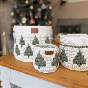 May include: Three white crochet baskets with green Christmas tree patterns. Each basket features a brown leather tag. The baskets, of varying sizes, are displayed on a wooden table. A Christmas tree is visible in the background.