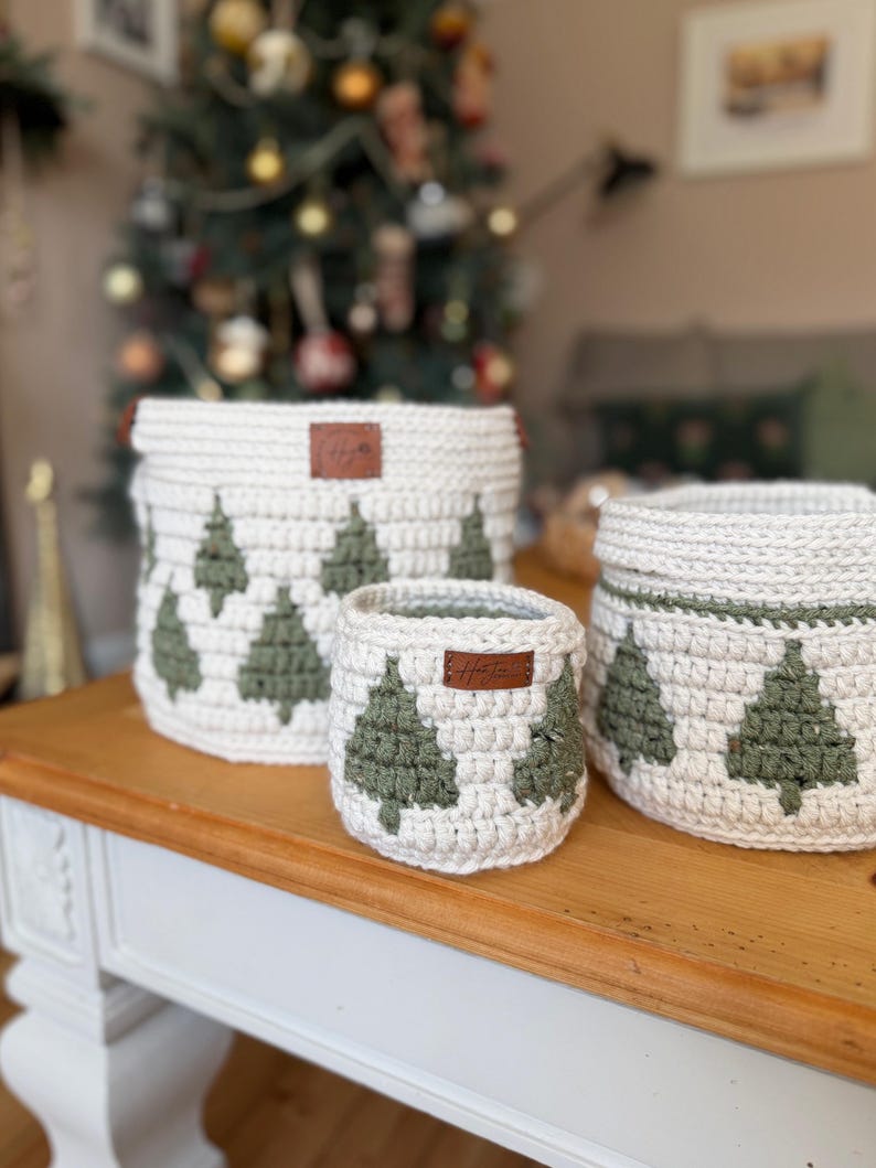 May include: Three white crochet baskets with green Christmas tree patterns. Each basket features a brown leather tag. The baskets, of varying sizes, are displayed on a wooden table. A Christmas tree is visible in the background.