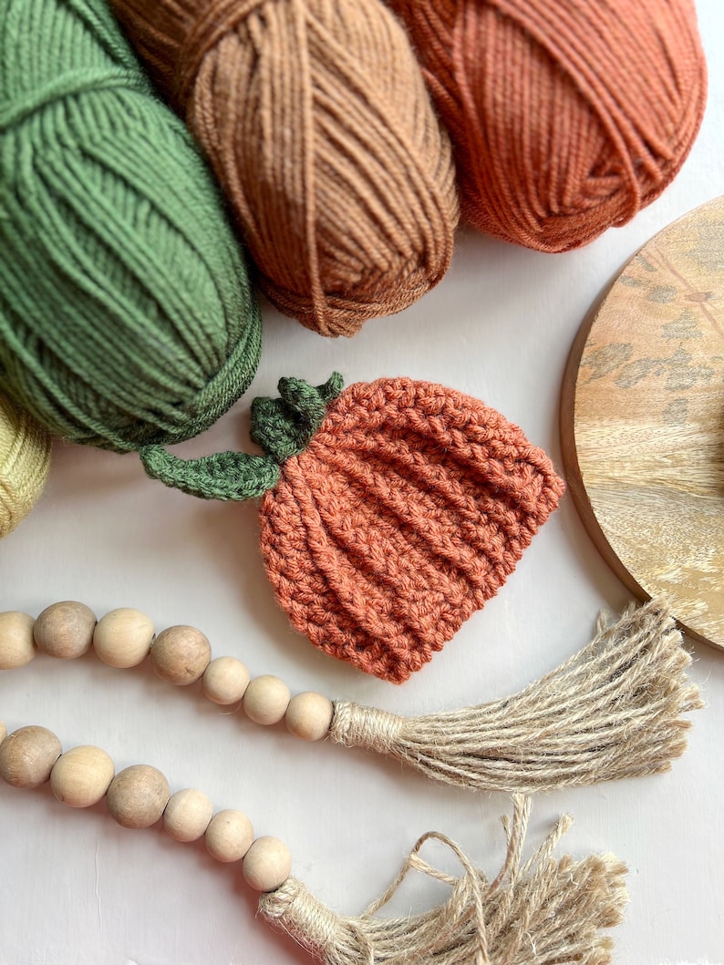 May include: A crocheted orange pumpkin with green leaves, yarn balls in green, brown, and orange, and a wooden bead garland with a jute tassel.