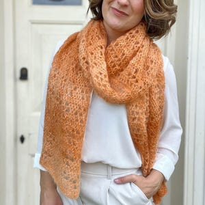 May include: A woman wearing a white shirt and beige trousers is wearing a crocheted scarf in a light orange colour. The scarf has a delicate, lacy pattern.