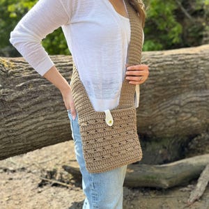 May include: A tan crochet crossbody bag with a long strap and a white leather closure. The bag has a textured pattern and is worn over the shoulder. The person is wearing a white cardigan and blue jeans.