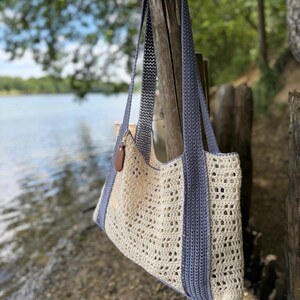 Puede incluir: Un bolso tote de ganchillo con un cuerpo calado de color crema y un patr&oacute;n texturizado. El bolso incluye dos correas largas de hombro en azul s&oacute;lido y un borde azul a los lados. Una peque&ntilde;a etiqueta de cuero marr&oacute;n en forma de gota cuelga.