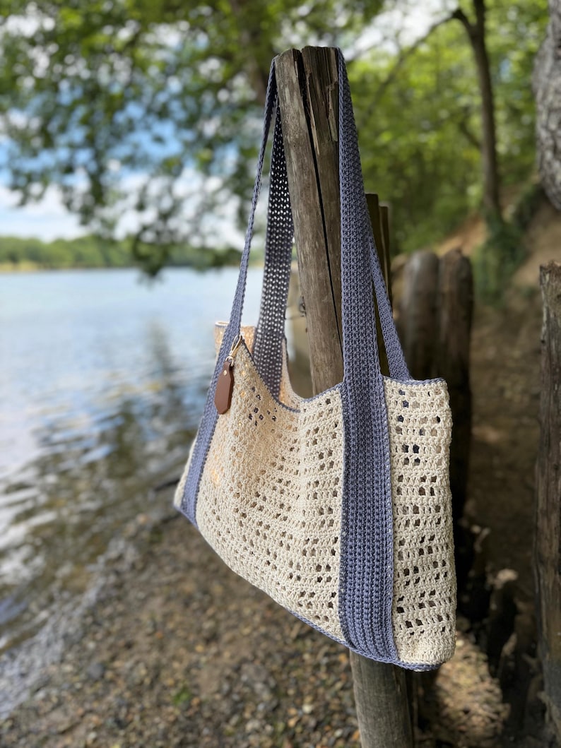 Puede incluir: Un bolso tote de ganchillo con un cuerpo calado de color crema y un patr&oacute;n texturizado. El bolso incluye dos correas largas de hombro en azul s&oacute;lido y un borde azul a los lados. Una peque&ntilde;a etiqueta de cuero marr&oacute;n en forma de gota cuelga.