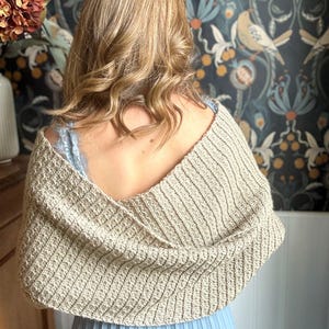 May include: A beige crocheted shawl with a v-neck design. The shawl is draped over the shoulders of a person wearing a blue dress.