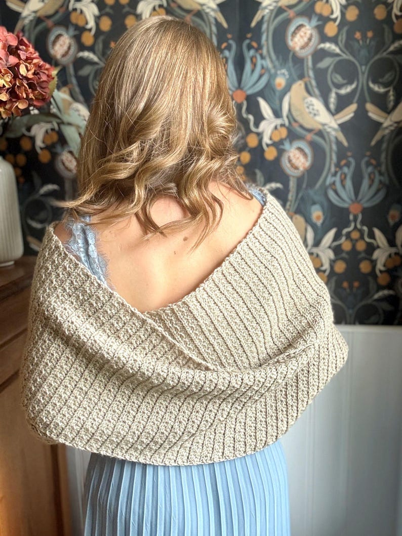 May include: A beige crocheted shawl with a v-neck design. The shawl is draped over the shoulders of a person wearing a blue dress.