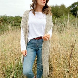 May include: A woman wearing a long, beige crochet cardigan over a white t-shirt and blue jeans. She is standing in a field of tall grass.