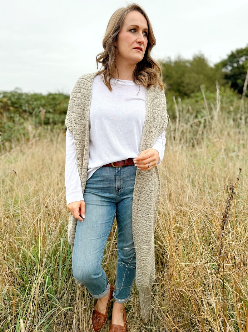 May include: A woman wearing a long, beige crochet cardigan over a white t-shirt and blue jeans. She is standing in a field of tall grass.