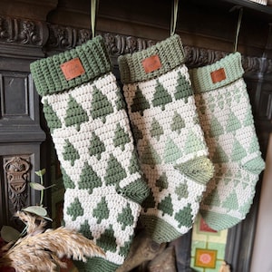 Puede incluir: Tres calcetines de Navidad hechos a mano con un patrón de árboles verdes y blancos. Cada calcetín tiene un puño verde y una pequeña etiqueta de cuero. Los calcetines cuelgan de una repisa oscura con cinta verde.