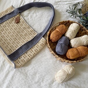 Puede incluir: Un bolso de ganchillo color crema con borde y correa de hombro gris oscuro, que incluye una peque&ntilde;a etiqueta de cuero marr&oacute;n. Junto al bolso hay una cesta tejida con cinco ovillos de lana en crema, gris y marr&oacute;n, y un ovillo extra.