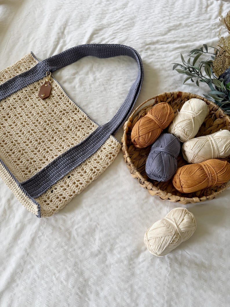 Puede incluir: Un bolso de ganchillo color crema con borde y correa de hombro gris oscuro, que incluye una peque&ntilde;a etiqueta de cuero marr&oacute;n. Junto al bolso hay una cesta tejida con cinco ovillos de lana en crema, gris y marr&oacute;n, y un ovillo extra.