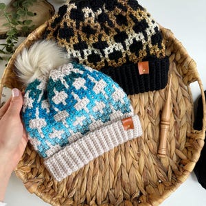 May include: Two crocheted hats with faux fur pom poms. One hat is black, brown, and white with a leopard print pattern. The other hat is blue, white, and beige with a geometric pattern.