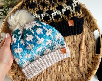 May include: Two crocheted hats with faux fur pom poms. One hat is black, brown, and white with a leopard print pattern. The other hat is blue, white, and beige with a geometric pattern.