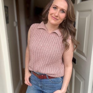 May include: A sleeveless, pale pink knitted top with a collared neckline. The top is worn with blue jeans and a brown belt. The outfit is worn in a doorway with natural light.