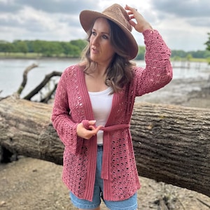 May include: A dusty rose crochet cardigan with a tie belt, worn over a white top and denim shorts. A straw hat is worn on the head. The cardigan features an open weave design.