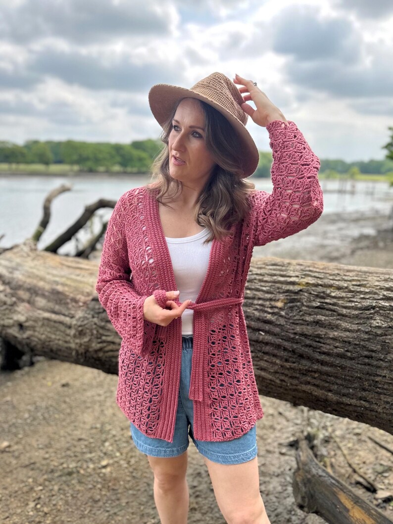 May include: A dusty rose crochet cardigan with a tie belt, worn over a white top and denim shorts. A straw hat is worn on the head. The cardigan features an open weave design.