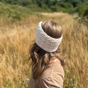 May include: A cream-colored knitted headband is worn by a person with brown hair. The headband is wide and textured, with a subtle pattern. The person is wearing a brown shirt and is standing in a field of tall, dry grass under a blue sky.