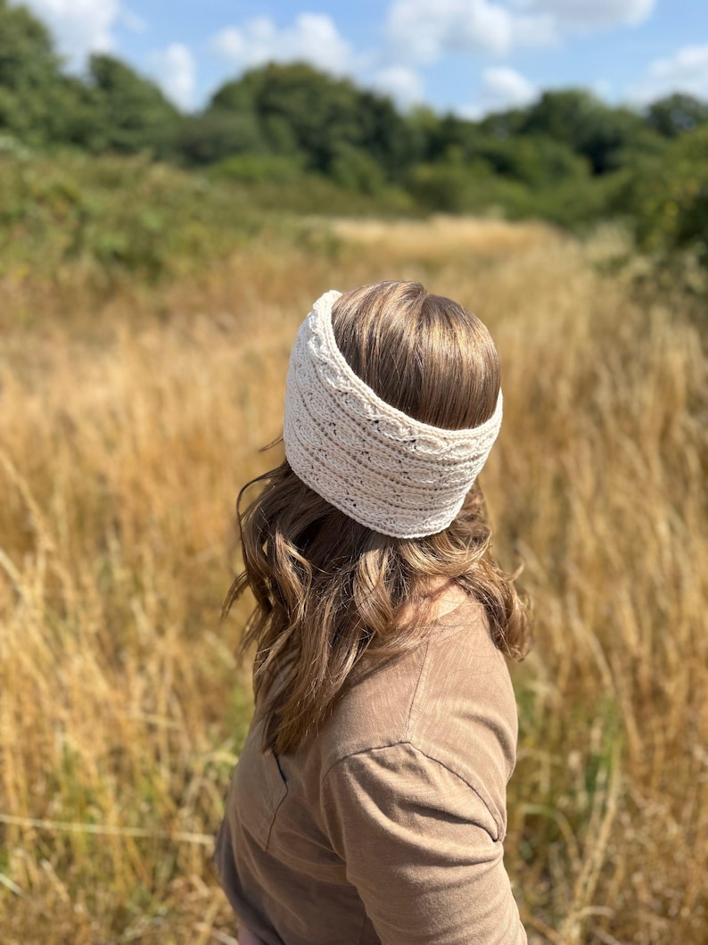 May include: A cream-colored knitted headband is worn by a person with brown hair. The headband is wide and textured, with a subtle pattern. The person is wearing a brown shirt and is standing in a field of tall, dry grass under a blue sky.