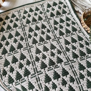 May include: A rectangular crochet blanket featuring a repeating grid pattern of dark forest green pine trees against a cream-colored background. The design is bordered by a solid dark green edge, showcasing a textured, hand-stitched yarn construction.